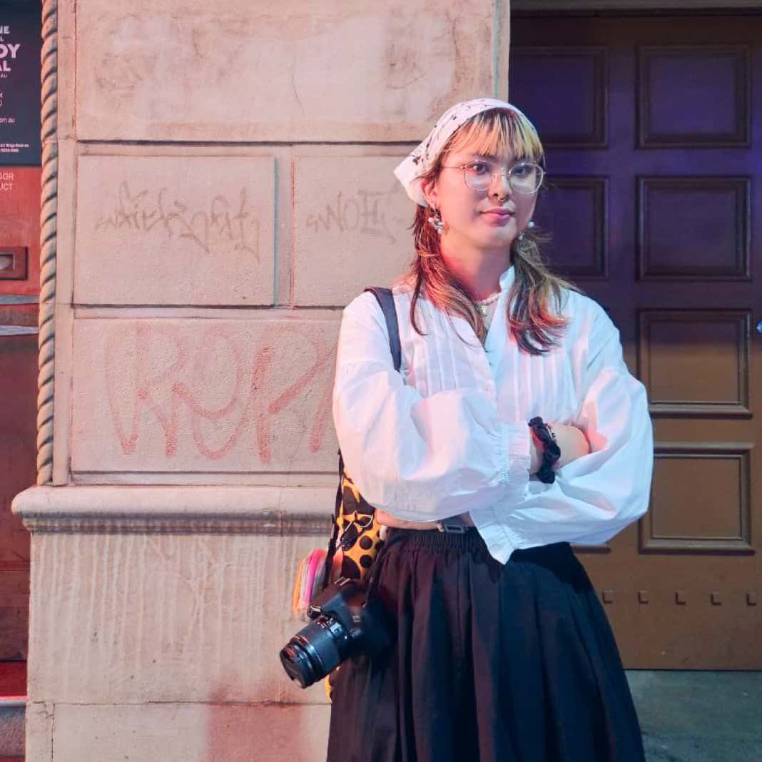 Portrait of Lauryn wearing a white shirt, glasses, and a printed headscarf, standing against an urban wall with faint graffiti lit by colorful street lights. They has a camera and a Yayoi Kusama tote bag slung at their side and a calm, composed expression.